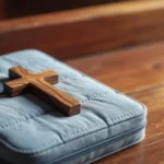 Primer plano de una pequeña cruz de madera pulida sobre una Biblia con funda acolchada azul claro y fondo bokeh.