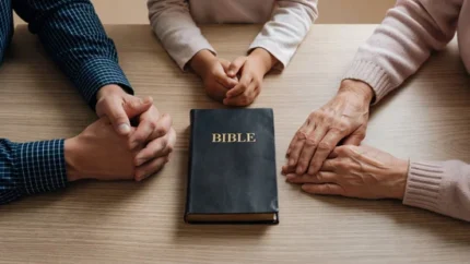 Manos de hombre, niño y anciana rodeando una Biblia negra con letras doradas sobre una mesa de madera clara.