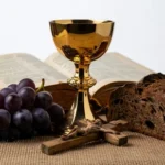 Cáliz dorado con pan artesanal roto, racimo de uvas moradas y biblia antigua sobre tela de yute y fondo blanco.