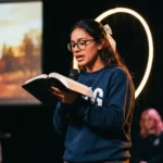 Joven mujer gafas moño blanco leyendo biblia abierta micrófono escenario oscuro aro luz circular cálida halo adoración contemporánea vertical desenfoque fondo