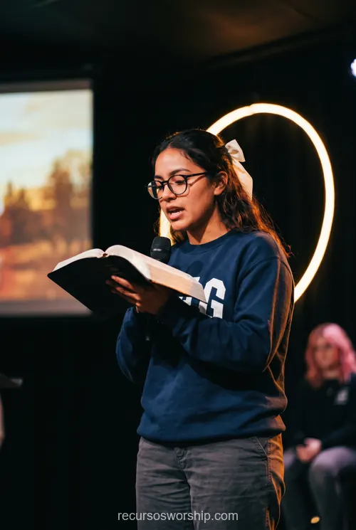 Joven mujer gafas moño blanco leyendo biblia abierta micrófono escenario oscuro aro luz circular cálida halo adoración contemporánea vertical desenfoque fondo