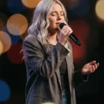 Mujer rubia cantando ojos cerrados microfono blazer gris fondo oscuro luces bokeh calidas naranja amarillo adoracion alabanza vertical cinematografica