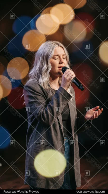 Mujer rubia cantando ojos cerrados microfono blazer gris fondo oscuro luces bokeh calidas naranja amarillo adoracion alabanza vertical cinematografica