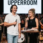 Pareja hombre mujer predicando microfonos camisa blanca falda oscura biblia abierta mesa madera pantalla UPPERROOM escenario iglesia banda bateria luces culto vertical luminoso
