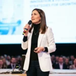 Mujer conferencista blazer blanco cuello negro microfono control remoto pantalla innovation future luces neon circulo purpura audiencia desenfocada escenario horizontal profesional