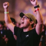 Joven hombre adorando brazos alzados gorra oliva gafas camiseta negra cantando pulseras multitud bokeh purpura naranja concierto cristiano vertical