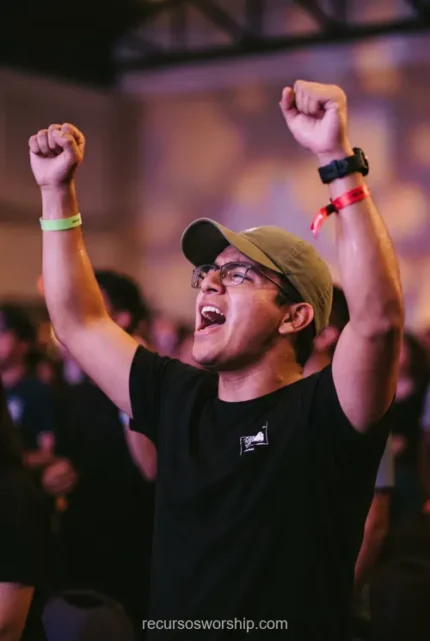 Joven hombre adorando brazos alzados gorra oliva gafas camiseta negra cantando pulseras multitud bokeh purpura naranja concierto cristiano vertical