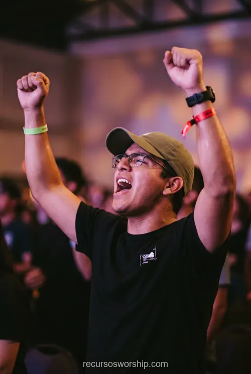 Joven hombre adorando brazos alzados gorra oliva gafas camiseta negra cantando pulseras multitud bokeh purpura naranja concierto cristiano vertical