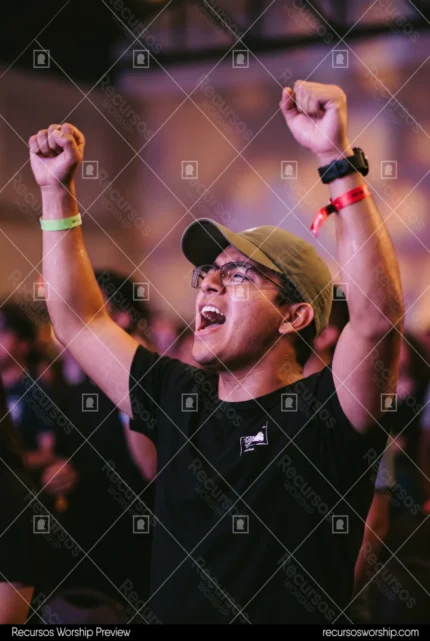 Joven hombre adorando brazos alzados gorra oliva gafas camiseta negra cantando pulseras multitud bokeh purpura naranja concierto cristiano vertical