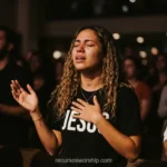 Mujer joven llorando lagrimas camiseta JESUS negro letras blancas mano pecho adoracion cabello rizado fondo multitud oscura bokeh cuadrado emotivo
