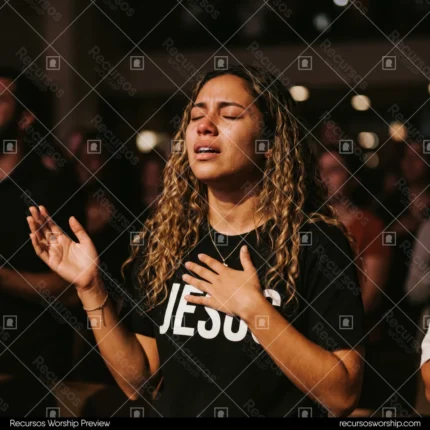 Mujer joven llorando lagrimas camiseta JESUS negro letras blancas mano pecho adoracion cabello rizado fondo multitud oscura bokeh cuadrado emotivo