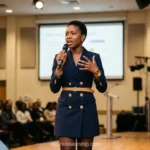 Mujer negra oradora blazer azul marino botones dorados cinturon camel microfono escenario madera audiencia pantalla joyas conferencia cuadrado profesional