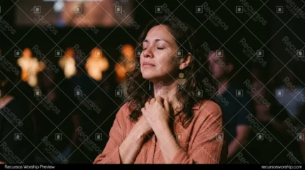 Mujer orando manos unidas pecho camisa terracota pendientes dorados ojos cerrados multitud culto luces calidas fondo oscuro horizontal intimo
