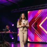 Mujer lider alabanza chaqueta denim beige microfono pantalla LED chevrones morado rosa teclado escenario luces culto contemporaneo vertical alegre