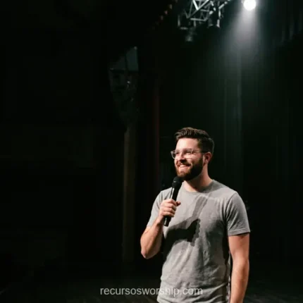Hombre joven predicando barba lentes camiseta gris microfono spotlight sombra escenario madera oscuro fondo negro sonrisa cuadrado minimalista
