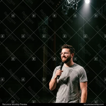 Hombre joven predicando barba lentes camiseta gris microfono spotlight sombra escenario madera oscuro fondo negro sonrisa cuadrado minimalista