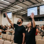 Pareja hombre mujer adorando manos alzadas ojos cerrados camisas negras congregacion pantalla SENHOR TU ES DIGNO iglesia moderna techo industrial cuadrado luminoso
