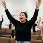 Mujer adoracion brazos alzados ojos cerrados sueter negro jeans sillas marrones congregacion iglesia vertical emotivo