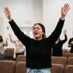 Mujer adoracion emocional brazos alzados ojos cerrados sueter negro jeans congregacion sillas marrones iglesia vertical