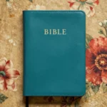 Biblia cerrada cubierta azul verdoso teal con inscripción dorada sobre papel tapiz vintage con flores rojas y hojas verdes cenital