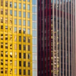 Fachada rascacielos modernos plano detalle vertical ventanas vidrio doradas amarillas azules reflejando entorno urbano franjas verticales rojas marron geometrica arquitectonica corporativa moderna textura