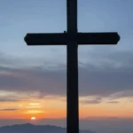 Cruz madera grande silueta oscura cima roca montanosa atardecer sol descendiendo montanas brumosas cielo azul gris degradado vertical contraluz solemne contemplativo viernes santo
