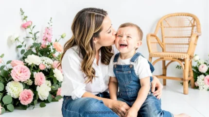 Madre morena cabello largo besando mejilla bebe varon riendo carcajadas estudio blanco luminoso flores rosas blancas silla ratan horizontal festivo