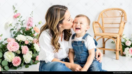 Madre morena cabello largo besando mejilla bebe varon riendo carcajadas estudio blanco luminoso flores rosas blancas silla ratan horizontal festivo