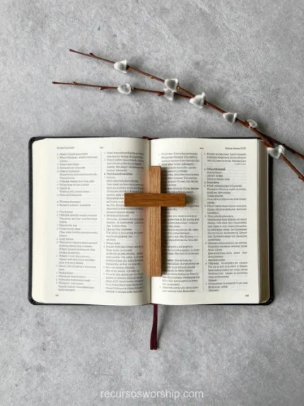 Biblia abierta en doble página con cruz de madera natural en el centro y rama seca con botones blancos sobre concreto gris cenital
