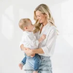 Madre joven rubia blusa blanca jeans sosteniendo bebe un año ropa blanca mirándola sonriendo fondo blanco puro estudio luminoso vertical plano americano