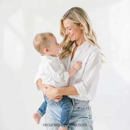 Madre joven rubia blusa blanca jeans sosteniendo bebe un año ropa blanca mirándola sonriendo fondo blanco puro estudio luminoso vertical plano americano