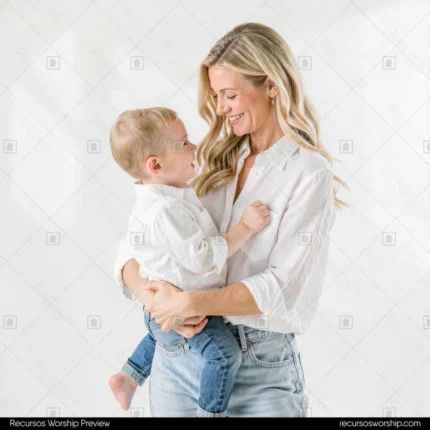 Madre joven rubia blusa blanca jeans sosteniendo bebe un año ropa blanca mirándola sonriendo fondo blanco puro estudio luminoso vertical plano americano