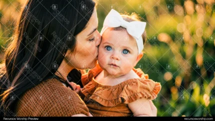 Madre morena cabello largo besando mejilla bebe niña ojos azules moño blanco vestido naranja exterior luz hora dorada bokeh verde horizontal cercano