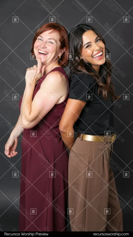 Madre pelirroja madura jumpsuit vino hija joven morena camiseta negra pantalón marron espalda espalda sonriendo riendo estudio fondo gris oscuro iluminación lateral vertical
