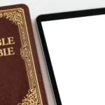 Biblia marrón rojizo con ornamentación dorada victoriana junto a tablet iPad pantalla blanca sobre fondo blanco liso vertical