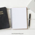Biblia negra Modern Ministry con letras doradas junto a cuaderno espiral rayado y bolígrafo con esquina de laptop sobre madera blanca cenital