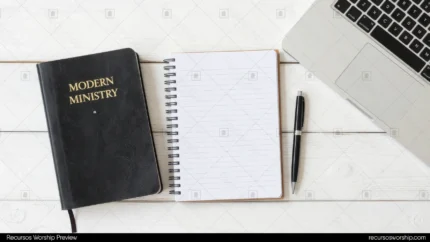 Biblia negra Modern Ministry con letras doradas junto a cuaderno espiral rayado y bolígrafo con esquina de laptop sobre madera blanca cenital