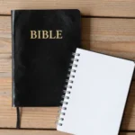 Biblia negra cerrada con letras doradas y marcapáginas rojo junto a cuaderno espiral abierto en blanco sobre madera clara natural cenital vertical