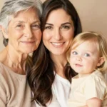 Abuela mayor cabello gris corto madre joven morena sonriendo bebe rubia ojos azules vestido amarillo tres generaciones estudio beige calido vertical plano busto sereno