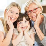 Abuela rubia gafas rojas madre joven rubia y niña pequeña morena apoyando mejillas en manos sonrisas amplias estudio luminoso calido vertical grupal