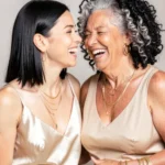 Madre mayor cabello cano rizado e hija joven morena riendo juntas estudio fondo beige vestidos dorados champan joyeria elegante vertical plano medio